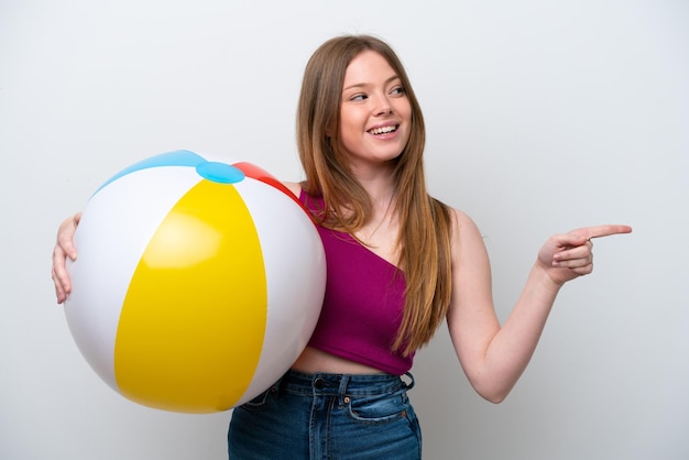 Jonge kaukasische vrouw die strandbal houdt die op witte achtergrond wordt geïsoleerd en met de vinger naar de zijkant wijst en een product presenteert