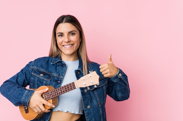 Jonge Kaukasische vrouw die ukelele speelt die en duim omhoog opheft glimlacht