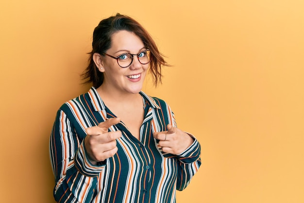 Jonge kaukasische vrouw die vrijetijdskleding en een bril draagt die met de vingers naar de camera wijst met een blij en grappig gezicht, goede energie en vibes