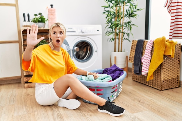 Jonge kaukasische vrouw die vuile was in de wasmachine doet en stopgebaar doet met boze handpalmen en frustratie-expressie