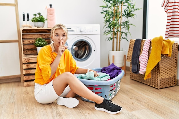 Jonge kaukasische vrouw die vuile was in de wasmachine stopt en vraagt om stil te zijn met de vinger op de lippen, stilte en geheim concept