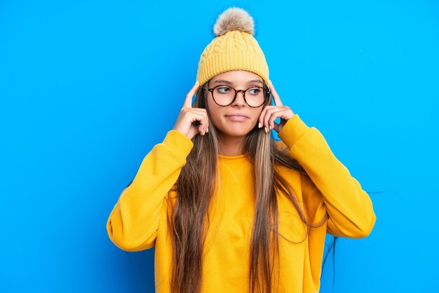 Jonge kaukasische vrouw die winterkleren draagt die op blauwe achtergrond worden geïsoleerd die twijfels hebben en denken