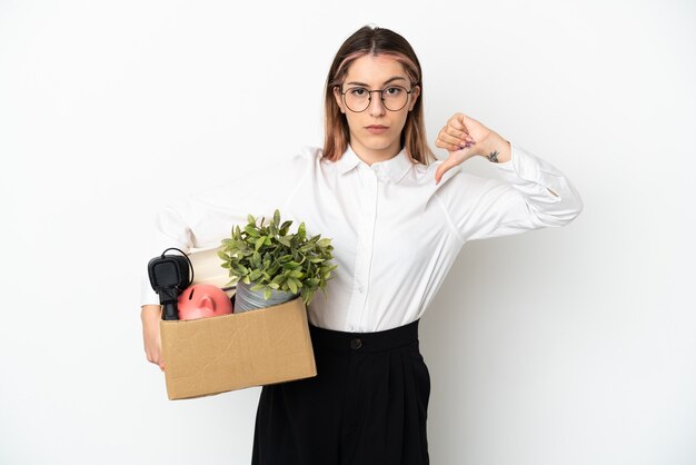 Jonge kaukasische vrouw die zich in nieuw huis onder dozen beweegt die op witte achtergrond worden geïsoleerd die duim met negatieve uitdrukking toont