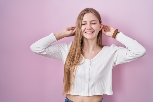 Jonge kaukasische vrouw die zich over roze achtergrond bevindt glimlachend die oren trekt met vingers grappig gebaar auditieprobleem