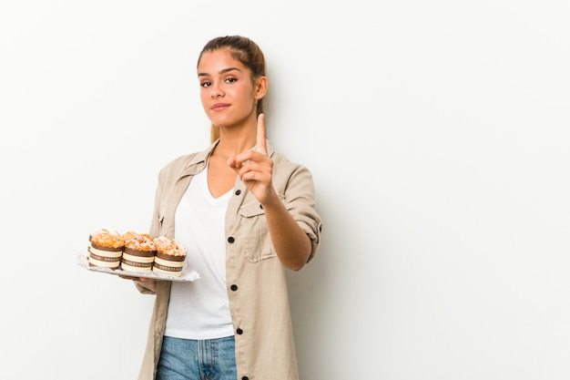 Jonge Kaukasische vrouw die zoete cakes houdt die nummer één met vinger tonen.