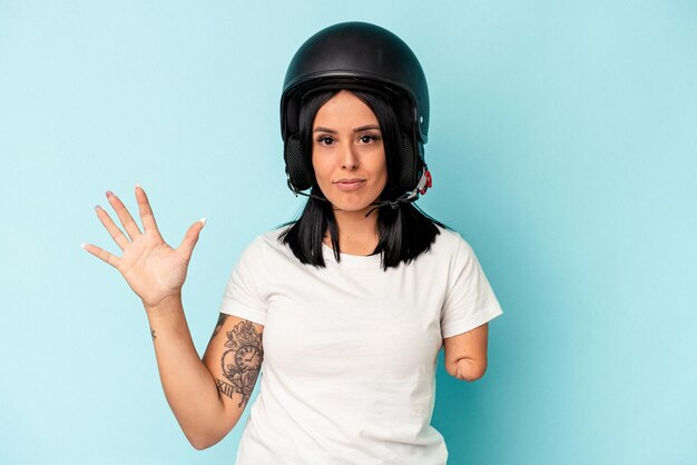 Jonge kaukasische vrouw met één arm die een motorhelm draagt, geïsoleerd op een blauwe achtergrond die vrolijk glimlacht en nummer vijf met vingers toont.