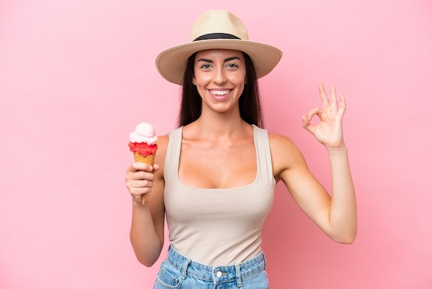 Jonge kaukasische vrouw met een cornetroomijs dat op roze achtergrond wordt geïsoleerd die ok teken met vingers toont