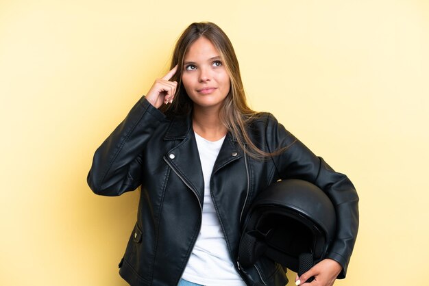 Jonge kaukasische vrouw met een motorhelm die op gele achtergrond wordt geïsoleerd die twijfels heeft en denkt