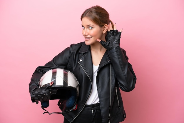 Jonge kaukasische vrouw met een motorhelm die op roze achtergrond wordt geïsoleerd die aan iets luistert door hand op het oor te leggen