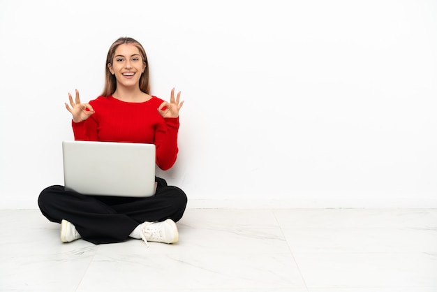 Jonge kaukasische vrouw met laptop die op de vloer zitten die een ok teken met vingers tonen