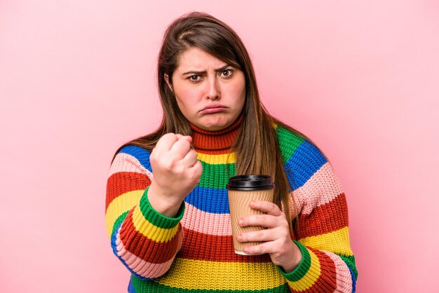 Foto jonge kaukasische vrouw met overgewicht die afhaalkoffie houdt geïsoleerd op roze achtergrond die vuist toont aan camera agressieve gezichtsuitdrukking