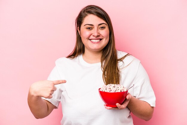 Jonge kaukasische vrouw met overgewicht die een kom ontbijtgranen houdt geïsoleerd op een roze achtergrondpersoon die met de hand wijst naar de ruimte van een shirtkopie, trots en zelfverzekerd