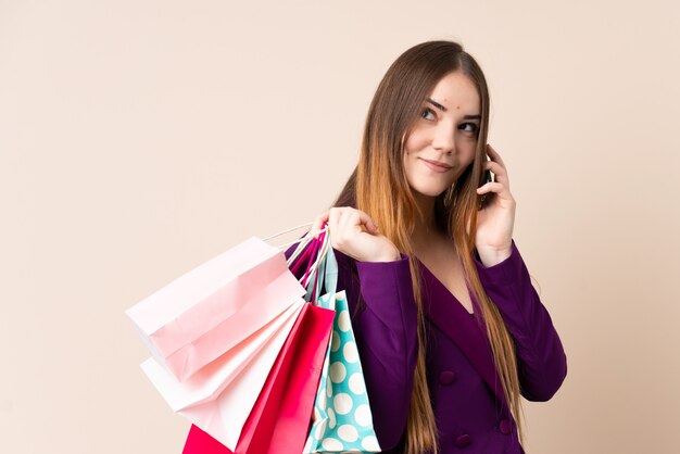 Jonge Kaukasische vrouw op beige muur met boodschappentassen en een vriend bellen met haar mobiele telefoon