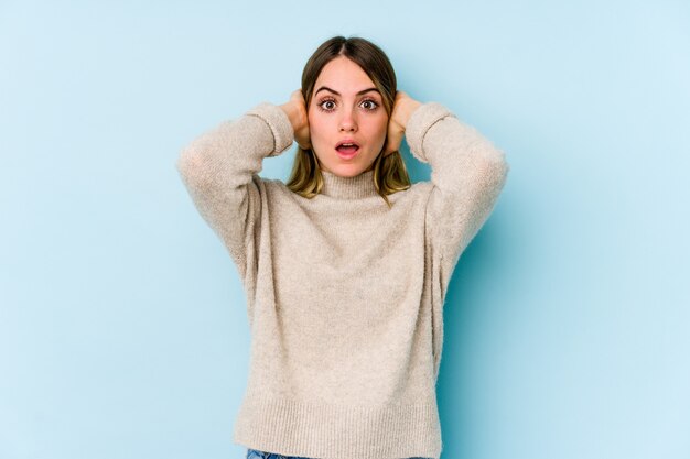 Jonge Kaukasische vrouw op blauwe muur die oren behandelt met handen die niet te hard geluid proberen te horen.