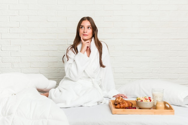 Jonge Kaukasische vrouw op het bed dat zijdelings met twijfelachtige en sceptische uitdrukking kijkt.