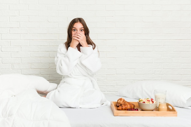 Jonge Kaukasische vrouw op het bed geschokt bedekkend mond met handen.