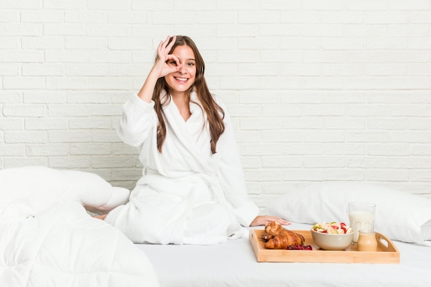 Jonge Kaukasische vrouw op het bed opgewonden het houden van ok gebaar op oog.