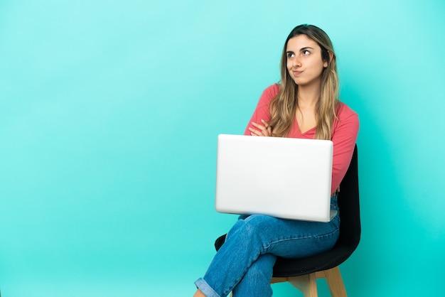 Jonge kaukasische vrouw zittend op een stoel met haar pc geïsoleerd op een blauwe achtergrond die twijfels maakt terwijl ze de schouders opheft