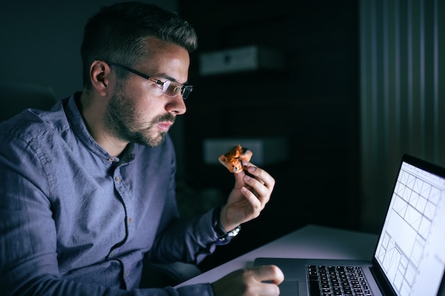 Jonge Kaukasische werknemer met oogglazen die snel voedsel eten en aan laptop werken terwijl het zitten laat op nacht in bureau.