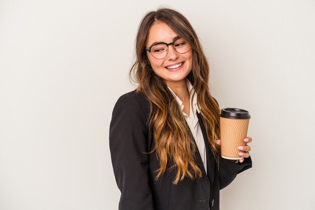 Jonge kaukasische zakenvrouw met een afhaalmaaltijdenkoffie geïsoleerd op een witte achtergrond kijkt opzij glimlachend, vrolijk en aangenaam.