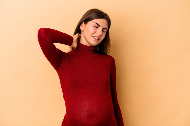 Jonge kaukasische zwangere vrouw die op beige muur wordt geïsoleerd die nekpijn lijdt als gevolg van een zittende levensstijl.