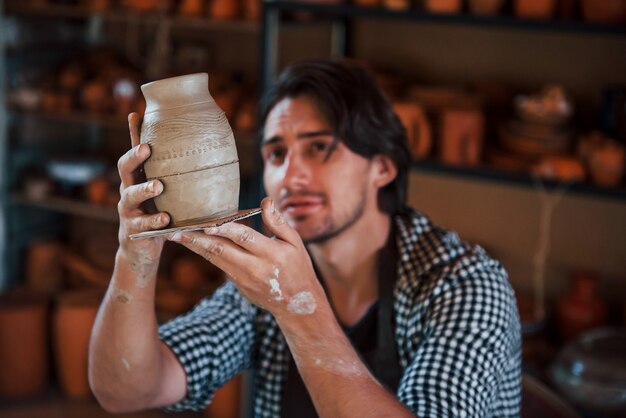 Jonge keramist houdt verse handgemaakte pot in de hand en kijkt naar de resultaten van zijn werk.