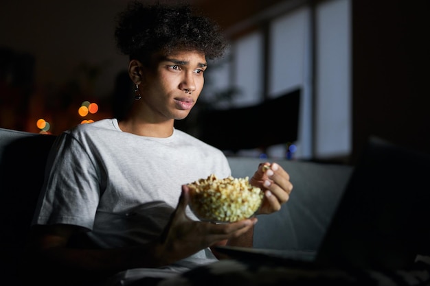 Jonge kerel die gefocust kijkt naar film online met laptop en popcorn eet terwijl hij op een stoel zit