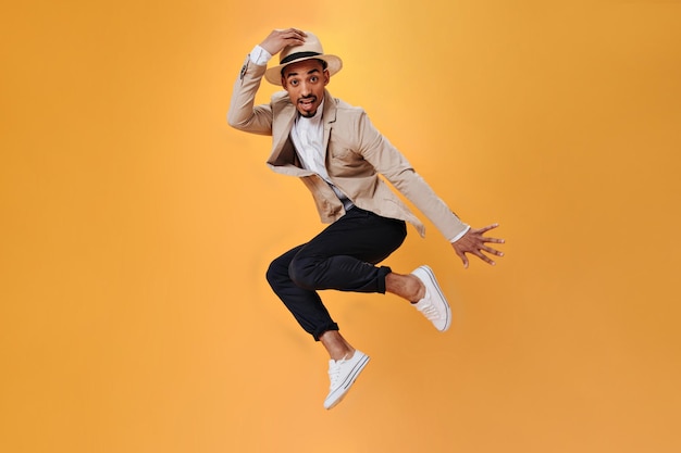 Jonge kerel in beige jas en hoed springt hoog op oranje achtergrond Portret van man in stijlvol pak poseren en dansen op geïsoleerde achtergrond