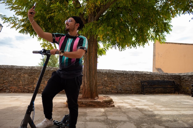 Jonge kerel met bril en een baard en vlechten in zijn haar die een selfie maken met zijn elektrische scooter