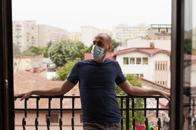 Jonge kerel met gezichtsmasker die op balkon ontspant tijdens coronavirusquarantaine.
