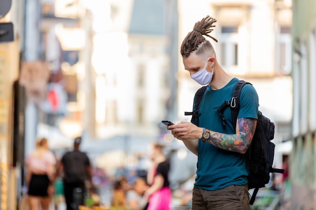 Jonge kerel met gezichtsmasker en dreadlocks gebruikt mobiele telefoon op straat