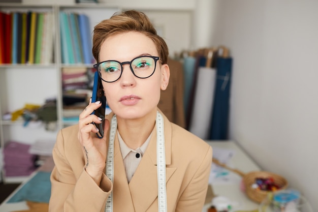 Jonge kleermaker in brillen praten op mobiele telefoon en bestelling opnemen tijdens het werken in de werkplaats