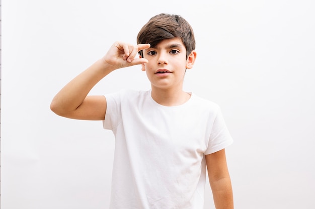 Jonge kleine jongen jongen dragen witte t-shirt staande over witte geïsoleerde achtergrond doen ok gebaar geschokt met verbaasd gezicht, oog kijken door vingers. Ongelovige uitdrukking.