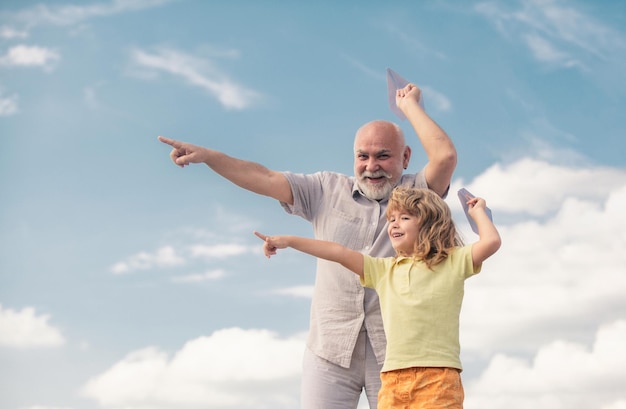 Jonge kleinzoon en oude grootvader spelen met een speelgoedpapiervliegtuig tegen de achtergrond van de zomerhemel