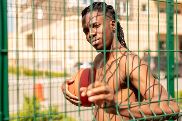 Jonge knappe Afrikaanse basketballer bal houden tijdens het kijken door hek bars omringende speelplaats op zonnige dag