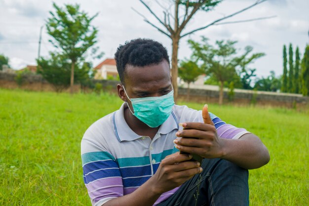 Jonge knappe afrikaanse man die buiten zit met een gezichtsmasker om te voorkomen, voorkomen, te voorkomen dat hij de uitbraak in de samenleving uitbreekt terwijl hij zijn mobiele telefoon bedient