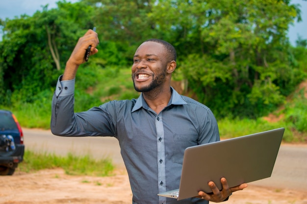 Jonge knappe afrikaanse zakenman die op een farnland staat en zich overenthousiast voelt terwijl hij zijn mobiel en laptop vasthoudt