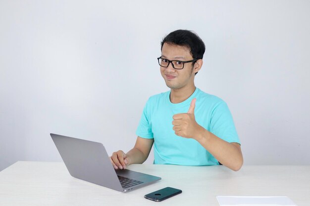 Jonge knappe aziatische man die duim opsteekt tijdens het werken met laptop Indonesische man met blauw shirt
