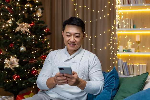 Jonge knappe aziatische man die thuis op de bank bij de kerstboom zit in een witte trui