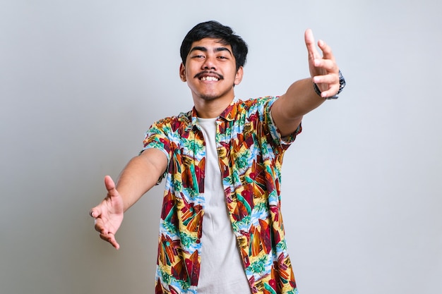 Jonge knappe aziatische man met een casual shirt kijkend naar de camera glimlachend met open armen voor een knuffel. vrolijke uitdrukking die geluk omhelst over witte achtergrond