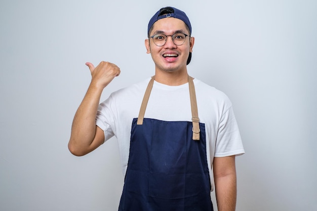 Jonge knappe barista-man met een schort met een grote glimlach op het gezicht wijzend met de hand en de vinger naar de zijkant kijkend naar de camera