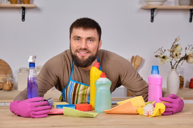 Jonge knappe bebaarde man in de keuken, laat al zijn schoonmaakpersoneel zien - wasmiddelen, borstels, sprays. Hij denkt dat hij klaar is voor de echte schoonmaak