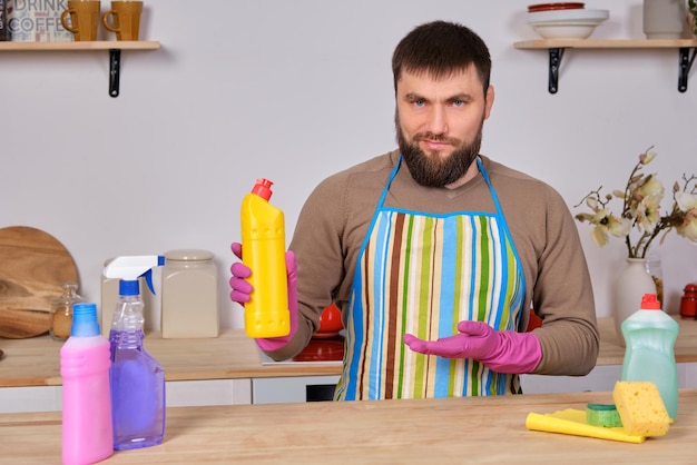 Jonge knappe bebaarde man in de keuken ruimt op met wasmiddelen, borstels, sprays