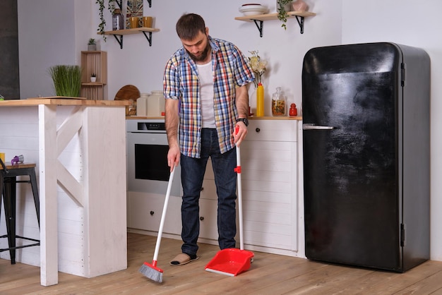 Jonge knappe bebaarde man in een keuken met een geruit hemd die de vloer veegt met een schop en b...