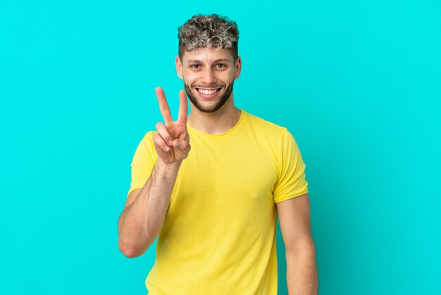 Jonge knappe blanke man geïsoleerd op blauwe achtergrond glimlachend en overwinningsteken tonen