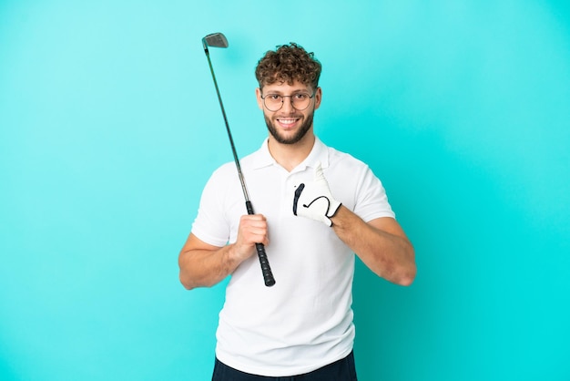 Jonge knappe blanke man geïsoleerd op blauwe achtergrond golfen en met duimen omhoog omdat er iets goeds is gebeurd