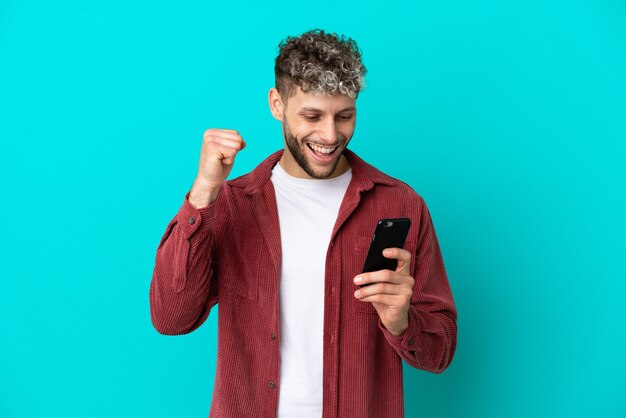 Jonge knappe blanke man geïsoleerd op blauwe achtergrond met behulp van mobiele telefoon en overwinningsgebaar doen