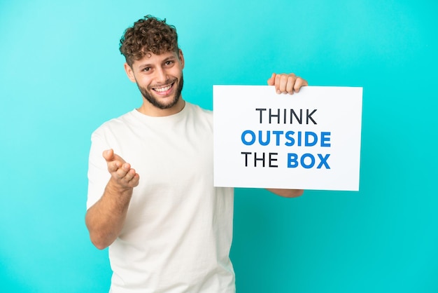 Jonge knappe blanke man geïsoleerd op blauwe achtergrond met een bordje met tekst Think Outside The Box die een deal maakt