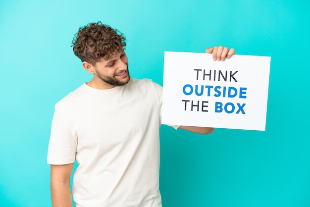 Jonge knappe blanke man geïsoleerd op blauwe achtergrond met een bordje met tekst Think Outside The Box