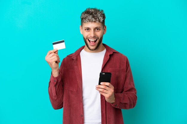 Jonge knappe blanke man geïsoleerd op een blauwe achtergrond die met de mobiel koopt en een creditcard vasthoudt met een verbaasde uitdrukking